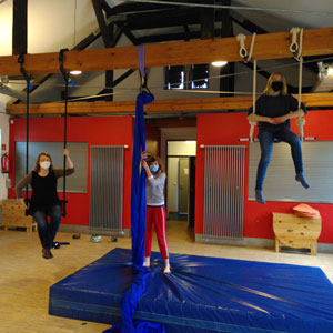 Drei Personen turnen an Luftakrobatikelementen, die an einem Balken im Saal befestigt sind.