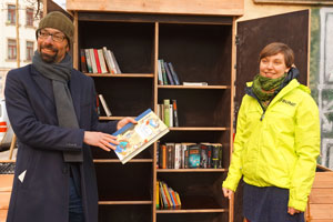 Zwei lächelnde, winterlich gekleidete Personen präsentieren ein Buch vor einem Bücherschrank im Feeien.