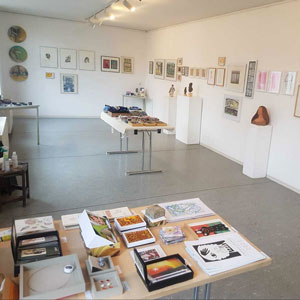 Blick in einen hellen Ausstellungsraum mit Grafiken an den Wänden und Objekten auf Tischen